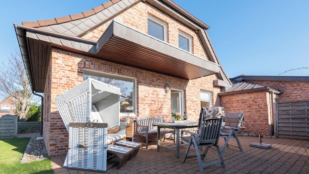 A two-story house with a brick exterior, a sloped roof, and a covered patio area with outdoor furniture. - Beautiful 2-room vacation apartment on the first floor with approx. 40 m² for up to 2 people (Westerland)