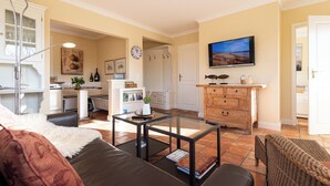 A living room with a sofa, coffee table, and a television. There is a kitchen in the background with cabinets and a clock. - Beautiful 2-room vacation apartment on the first floor with approx. 40 m² for up to 2 people (Westerland)