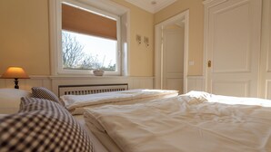 A bedroom with two beds, a window with blinds, a bedside lamp, and a radiator. - Beautiful 2-room vacation apartment on the first floor with approx. 40 m² for up to 2 people (Westerland)
