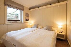 Een net opgemaakt bed met een witte deken en kussens, een nachtkastje met een lamp, een raam met uitzicht op groen en een plank met decoratieve items.
