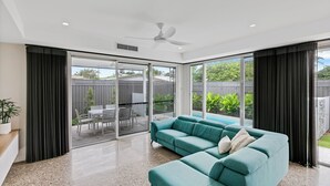 Ein modernes Wohnzimmer mit einem türkisfarbenen Sofa, Deckenventilator und großen Schiebetüren, die auf eine Terrasse im Freien führen.