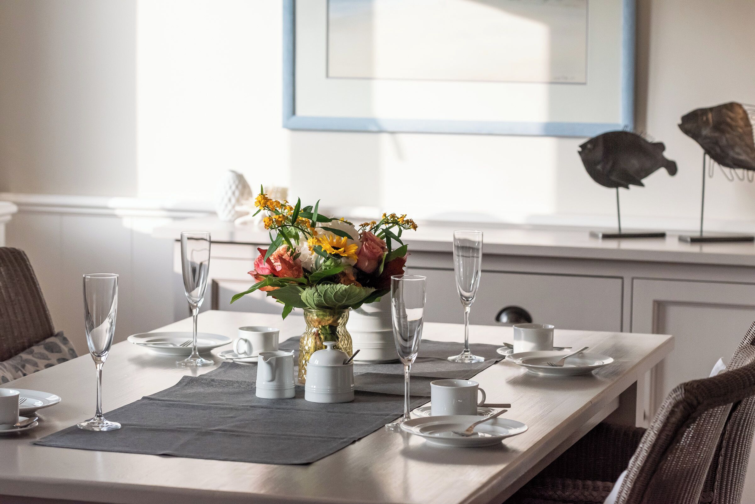 A dining table set with white plates, silverware, and clear glasses, accompanied by a vase of yellow flowers.