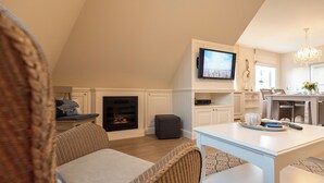 A cozy living room with a fireplace, a television mounted above it, and a dining area with a table and chairs. - Bright 3-room holiday apartment on approx. 100 m² for up to 4 people (Westerland)