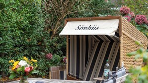 A cozy outdoor seating area with a wicker chair and a table, a sign reading "Söhuis," and a bottle of water on the table. - Bright 3-room holiday apartment on approx. 100 m² for up to 4 people (Westerland)