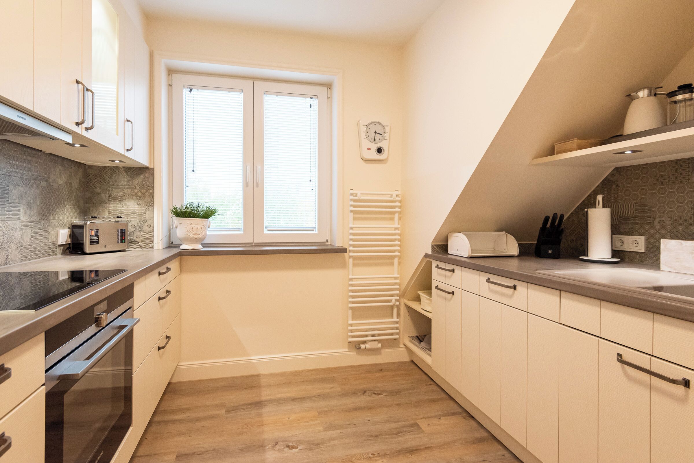 Une cuisine moderne équipée d’un four encastré, d’un four à micro-ondes et d’un grille-pain. On y trouve une fenêtre avec des stores, un porte-serviettes blanc et une horloge au mur.