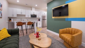 A modern kitchen with white cabinets, a microwave, and a refrigerator. There is a dining area with a table and chairs, and a TV mounted on the wall. - Trendy Tennyson Flat (Denver)