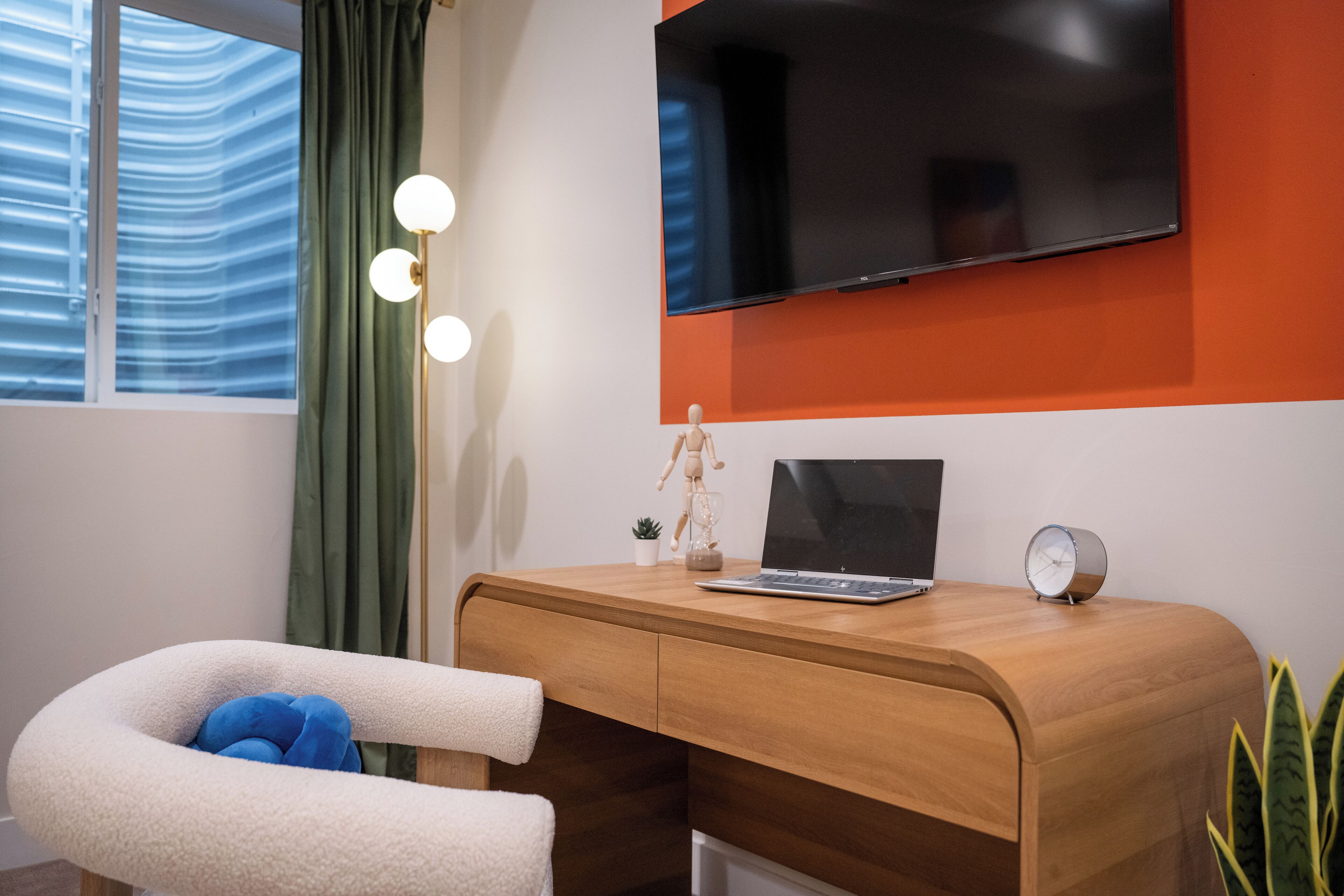 A modern room with a wooden desk, a laptop, a TV mounted on the wall, a small plant, and a chair with a cushion.