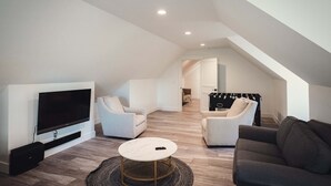 Una sala de estar moderna con una televisión de pantalla plana, dos sillones blancos, un sofá oscuro y una mesa de centro redonda.