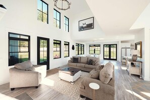 A spacious living room with a large sectional sofa, a coffee table, and a dining area with a table and chairs. There are multiple windows providing natural light and views of the outdoors.