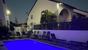 Una piscina iluminada con luces azules, rodeada de sillones de descanso y un edificio con ventanales arqueados.