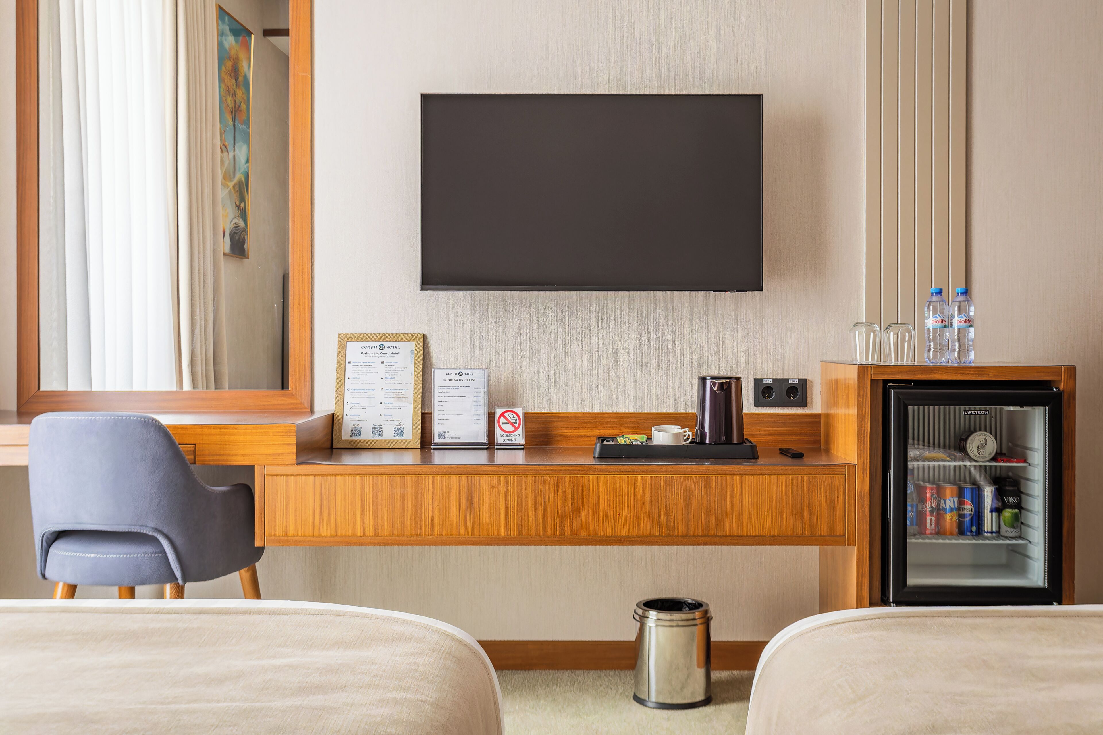 A hotel room with a bed, a desk, a television, a minibar, and a trash can.