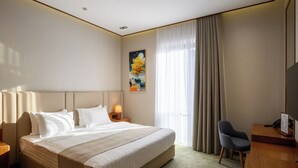 A hotel room with a large bed, a desk, a chair, a nightstand with a lamp, and a painting on the wall. - Consti Hotel Tashkent (Tashkent)