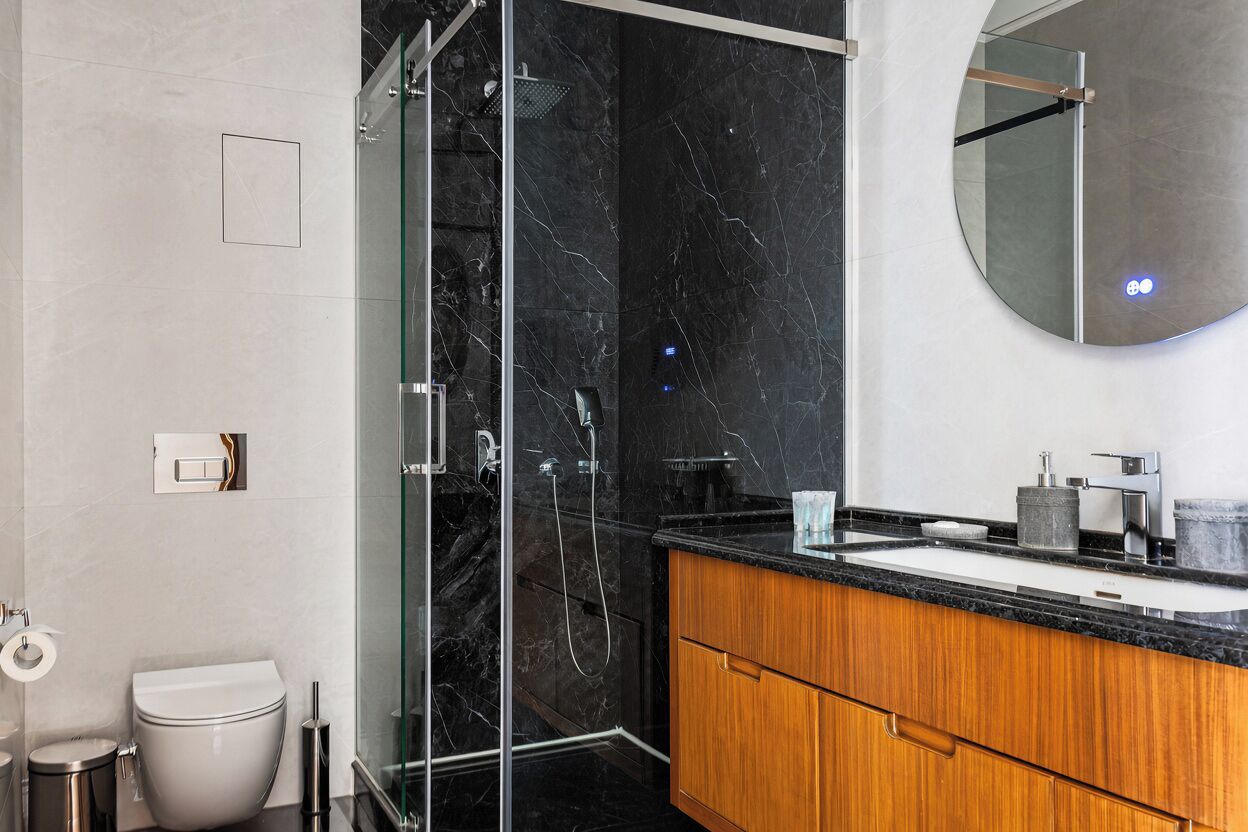 Een moderne badkamer met een douchecabine van glas, een marmeren wand, een houten wastafelmeubel en een ronde spiegel.