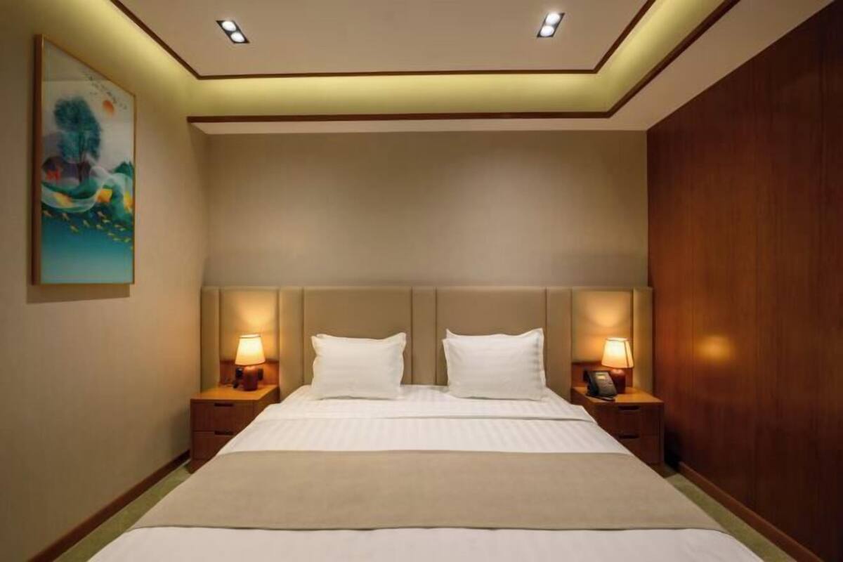 A hotel room with a large bed, two bedside tables with lamps, a painting on the wall, and recessed ceiling lighting.