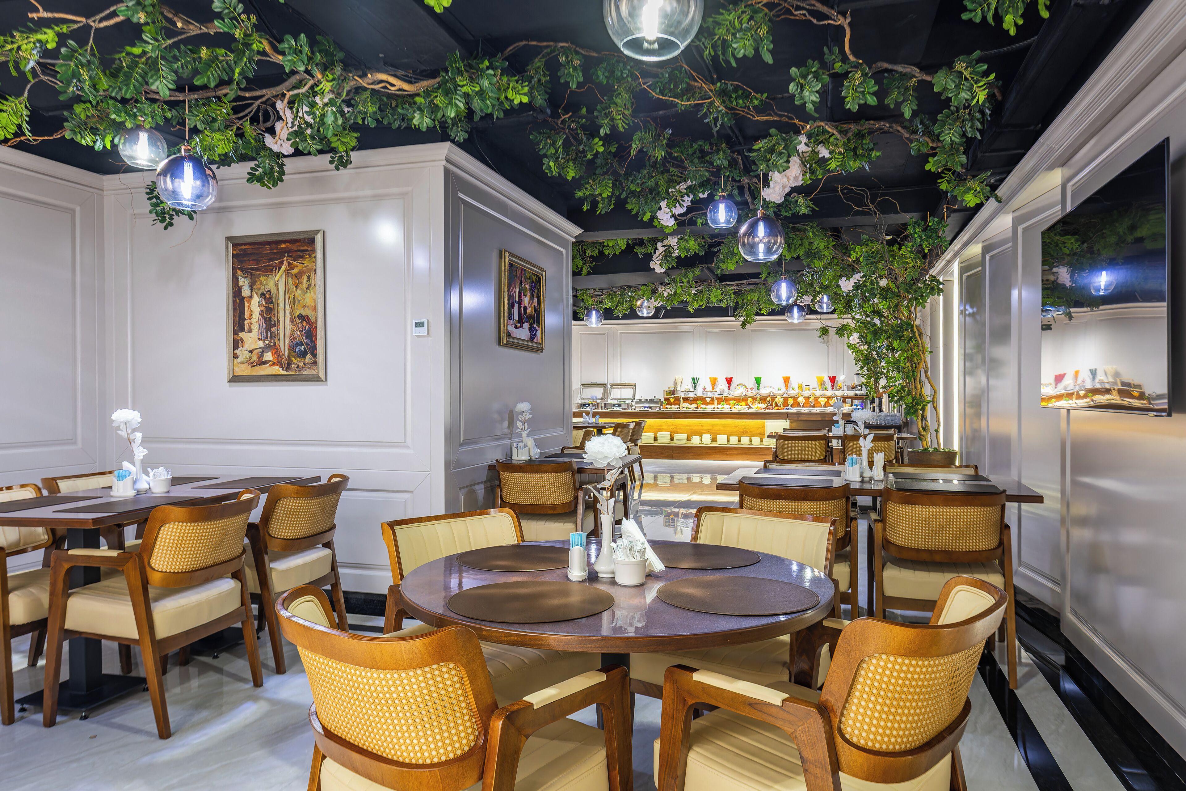A restaurant interior with round tables, wicker chairs, hanging plants, and pendant lighting.