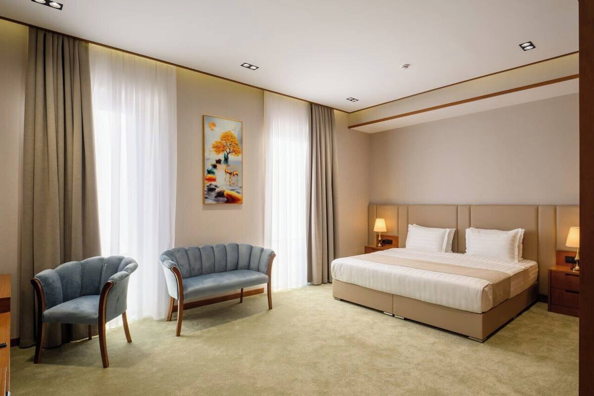 A hotel room with a large bed, two armchairs, a painting on the wall, and a window with curtains.