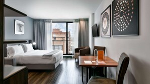 Ein modernes Hotelzimmer mit einem großen Bett, einem Schreibtisch mit Stuhl und einem Esstisch. Vor den Fenstern hängen Vorhänge und die Wände sind mit Kunstwerken geschmückt.