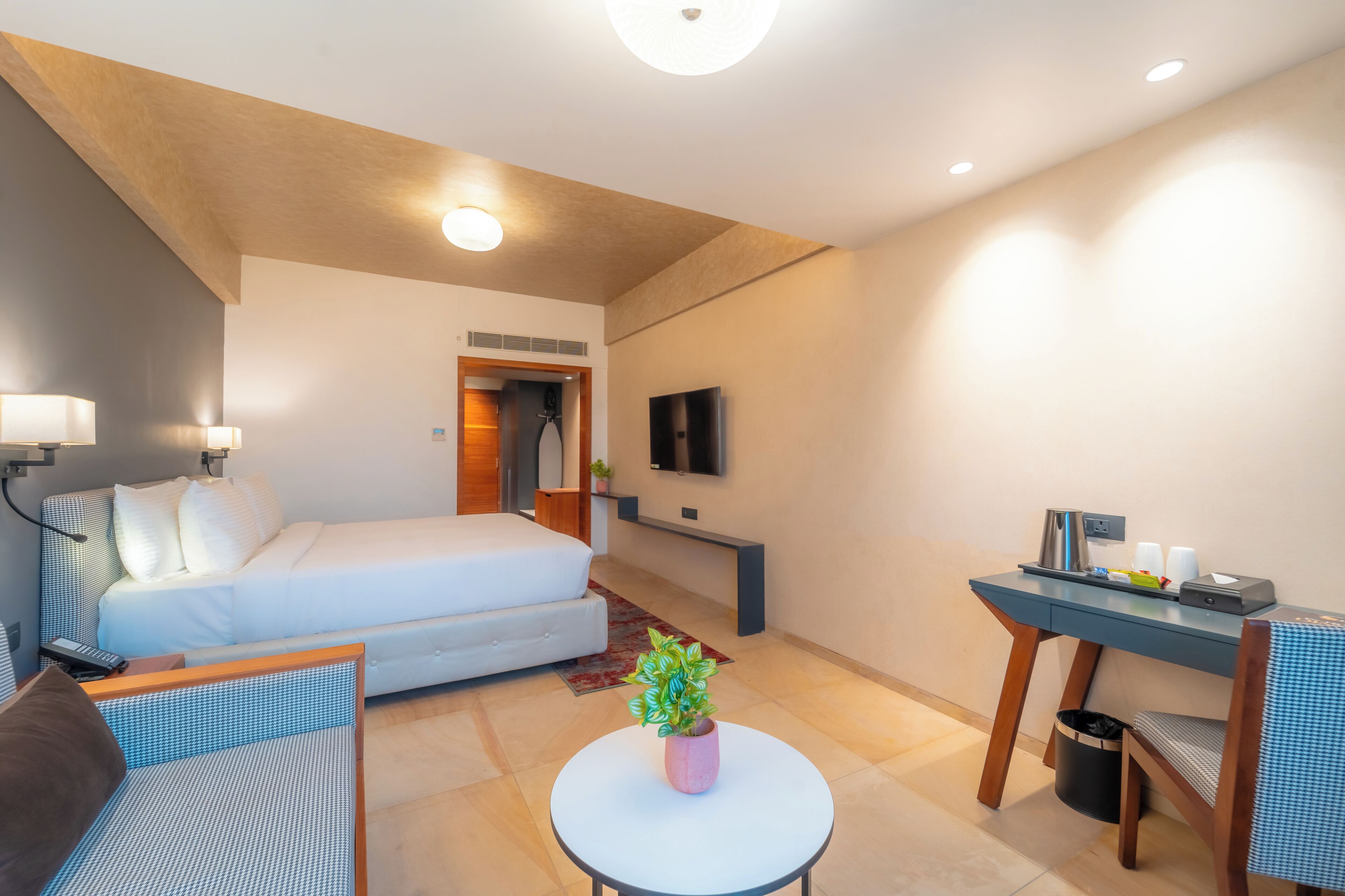 A modern hotel room with a bed, a desk, a chair, a small table with a plant, and a TV on the wall.