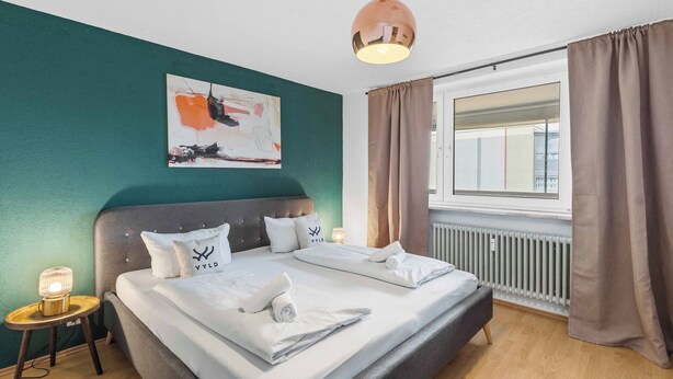 A neatly made bed with white linens and pillows, a bedside lamp, and a painting on the wall. - City Apartment | View & WIFI (Kempten (Allgäu))