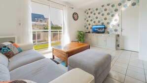 A modern living room with a sofa, ottoman, and a wooden coffee table. There is a television on a cabinet, and a window with a view of the outdoors. - Comfortable T3, walking distance to the beach - Secure garage (Dinard)