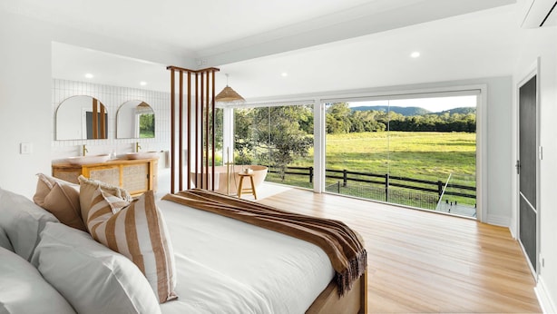 A bedroom with a large bed, a wooden dresser, a vanity, and a view of a green landscape through large windows. - Paddock View Ranch in the Sunny Coast Countryside (Belli Park)
