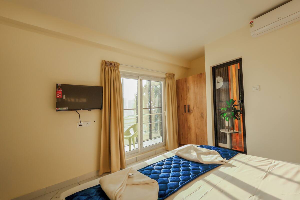 Une chambre à coucher avec un lit, un téléviseur fixé au mur, une fenêtre avec des rideaux et une porte en bois.