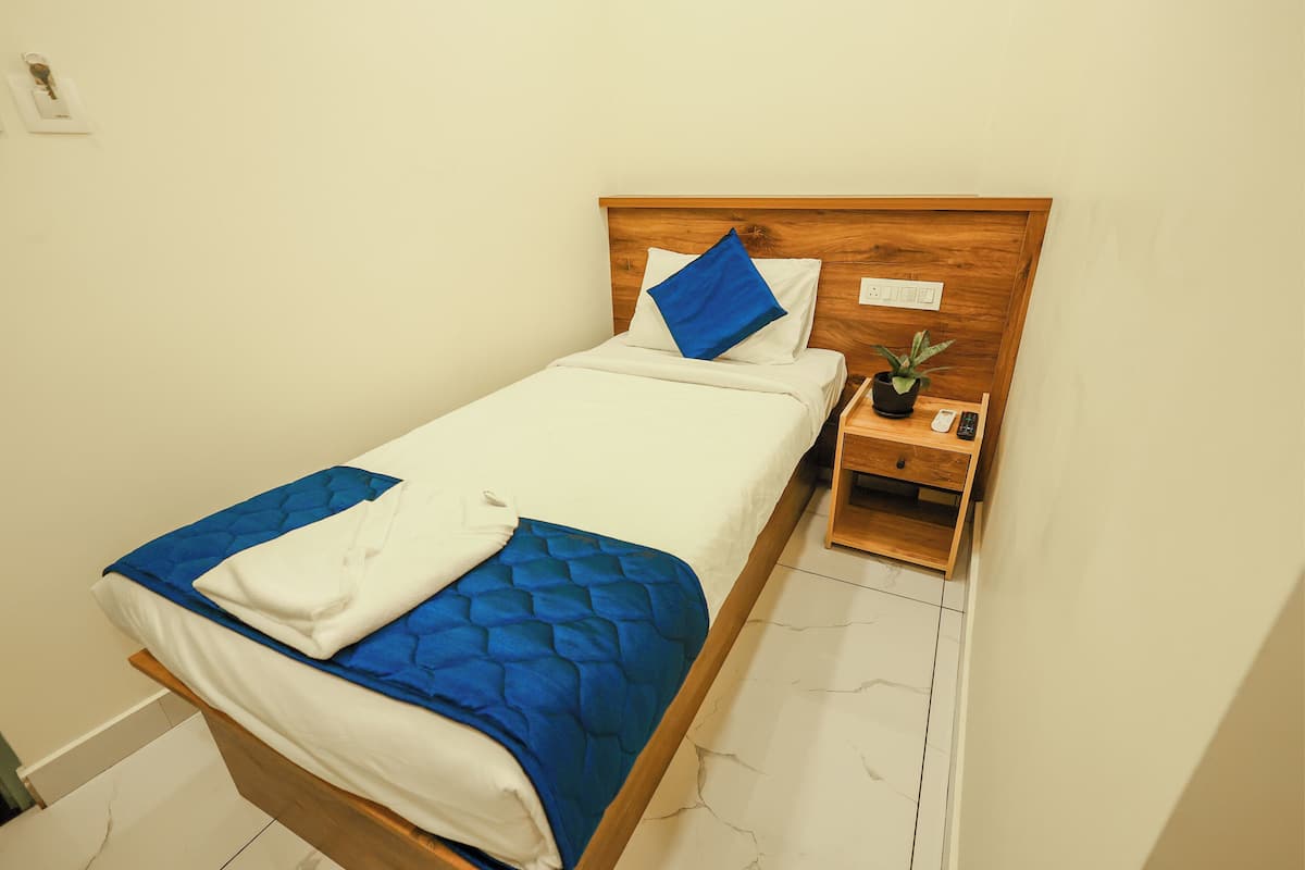 Un lit bien fait, recouvert d’une couette bleue, avec des oreillers blancs et un coussin bleu posé sur la tête de lit. Une petite table de chevet en bois, sur laquelle se trouvent une plante et une télécommande, est placée à côté du lit.