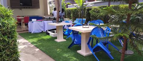 An outdoor dining area with tables and blue chairs, surrounded by greenery and a pathway.