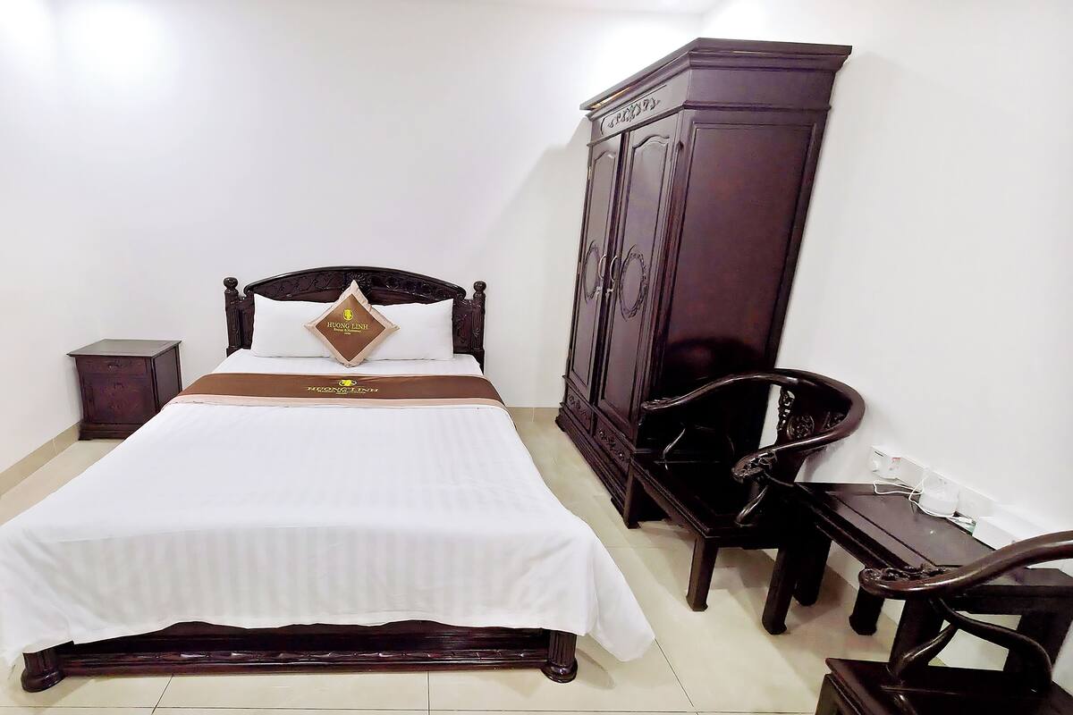 A neatly made bed with a headboard, a small bedside table, a dark wooden wardrobe, and a chair.