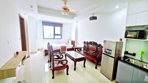 A hotel room with a bed, a dining table with chairs, a refrigerator, a microwave, a ceiling fan, and a window with blinds. - Huong Linh Apartment (Hanoi)