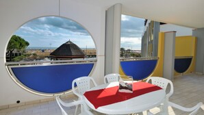 Ein Zimmer mit rundem Tisch und Stühlen, einer roten Tischdecke und Blick auf einen Strand mit einem Gebäude mit Reetdach.