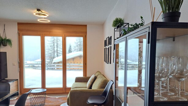 A modern living room with a sofa, chairs, a glass cabinet with wine glasses, and a view of a snowy landscape through large sliding doors.