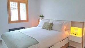 A bedroom with a bed, a bedside table with a lamp, a window, and a plant.