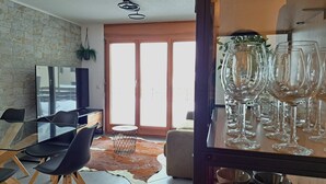 A modern living room with a glass dining table, black chairs, a TV, and a large window with a view of the outdoors.