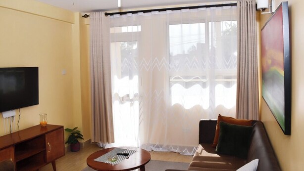 A living room with a flat-screen TV, a sofa, a coffee table, and a large window with sheer curtains.