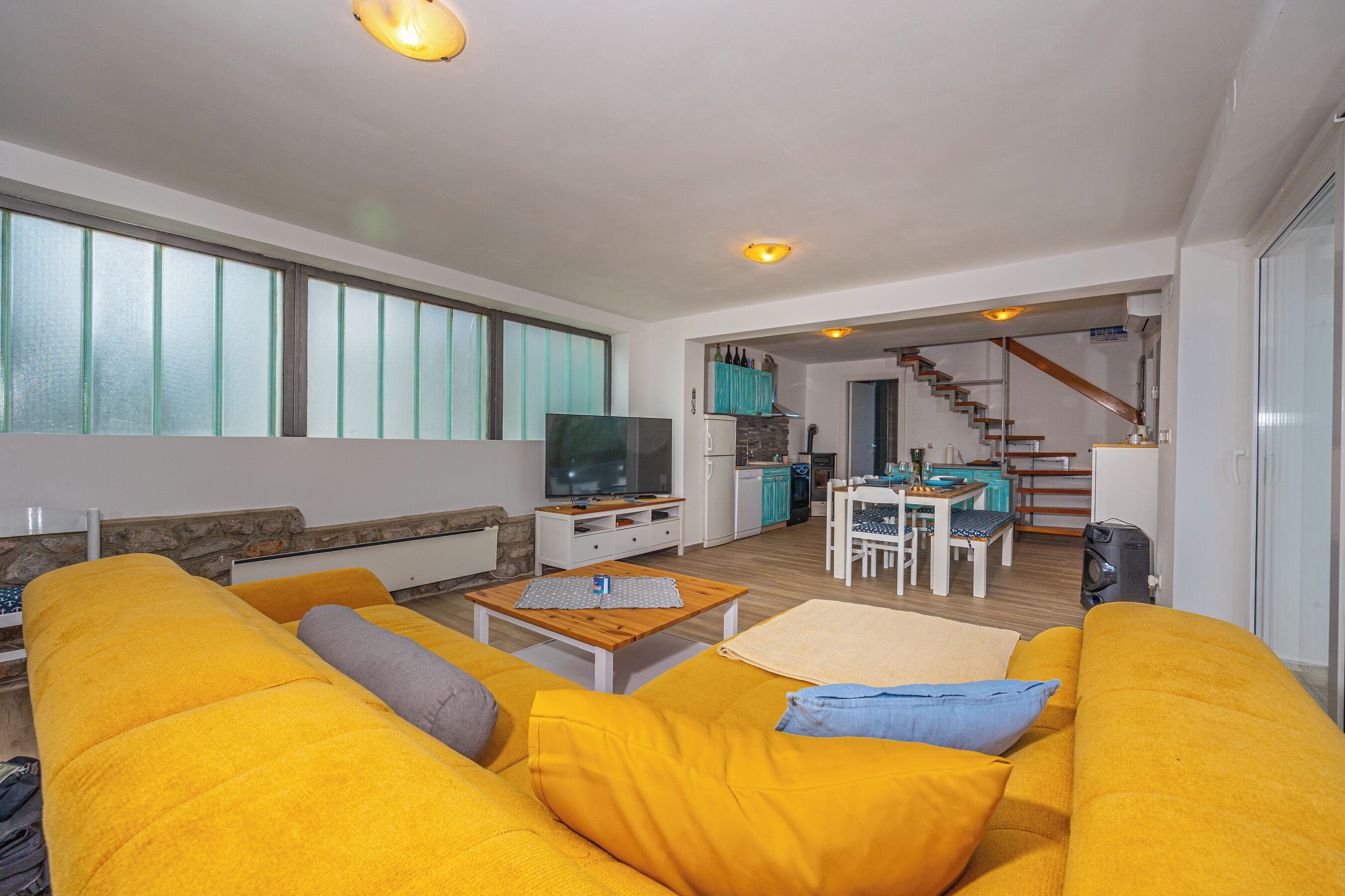 A bright living room with a yellow sofa, a wooden coffee table, and a TV. There is a kitchen in the background with a dining area and a staircase.