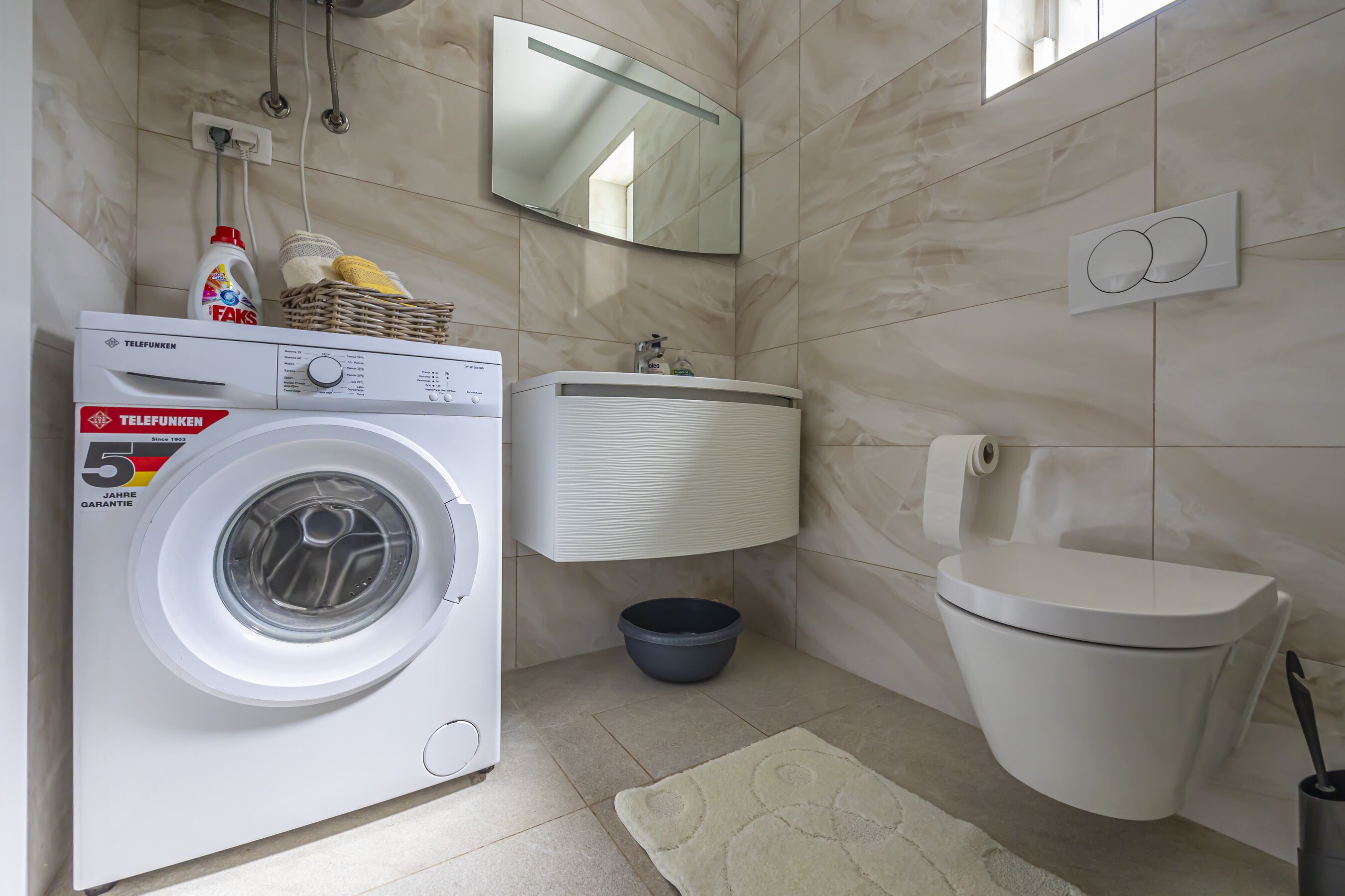 Et lille badeværelse med vaskemaskine, toilet, håndvask og spejl.