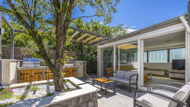 A modern outdoor living area with a barbecue, seating, and a glass-enclosed space with a TV. - Villa Barbara (Klenovica)