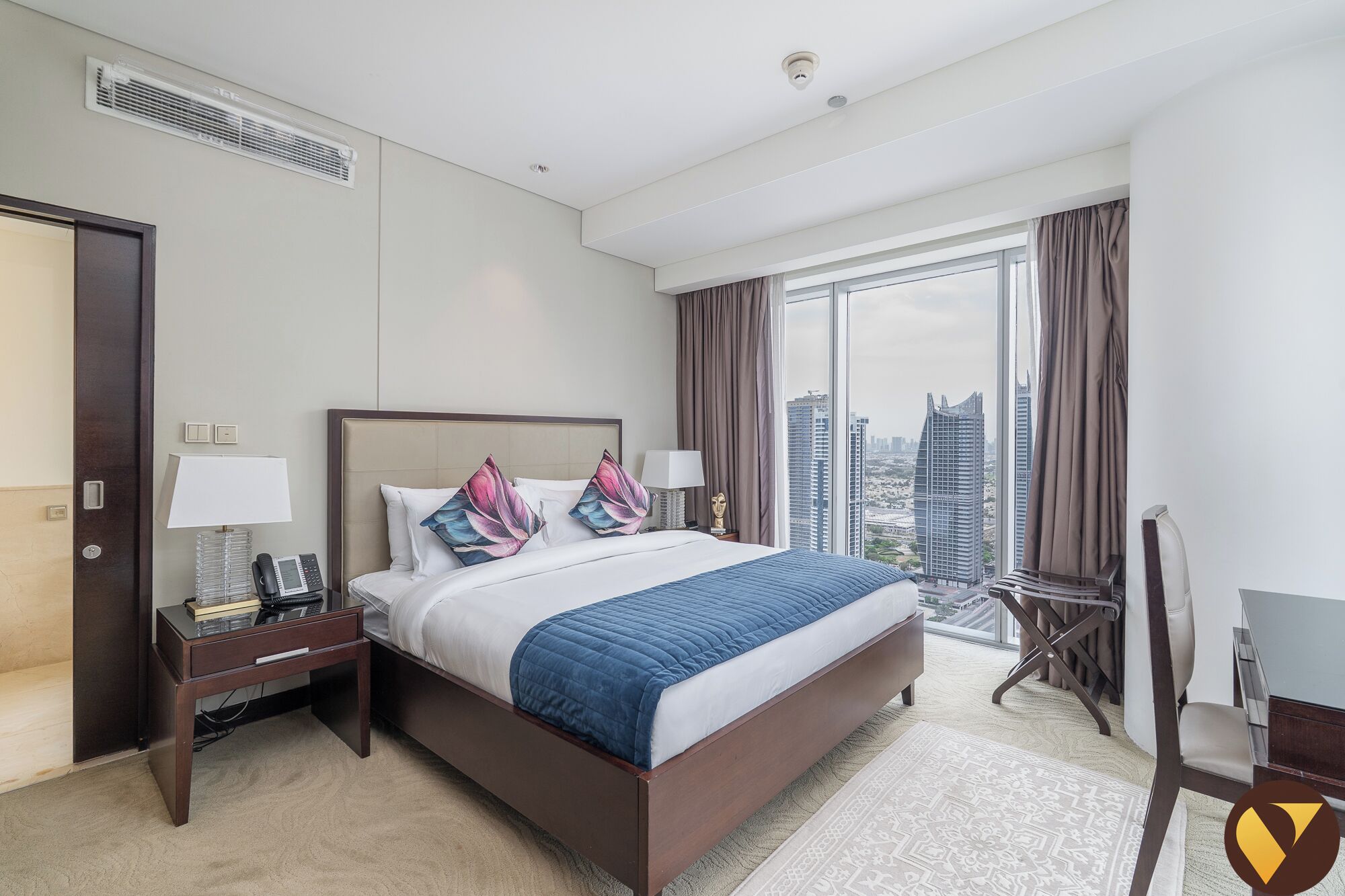 A hotel room with a large bed, a desk with a lamp, a chair, and a view of the cityscape.