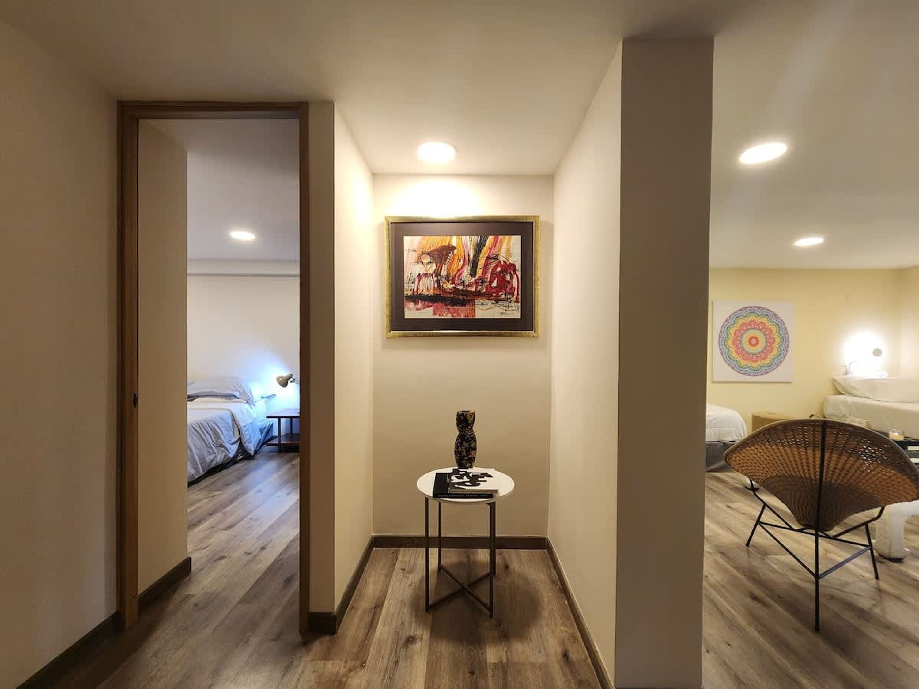 A hotel hallway with a wooden floor, a framed abstract painting, a small table with a decorative item, and a wicker chair.