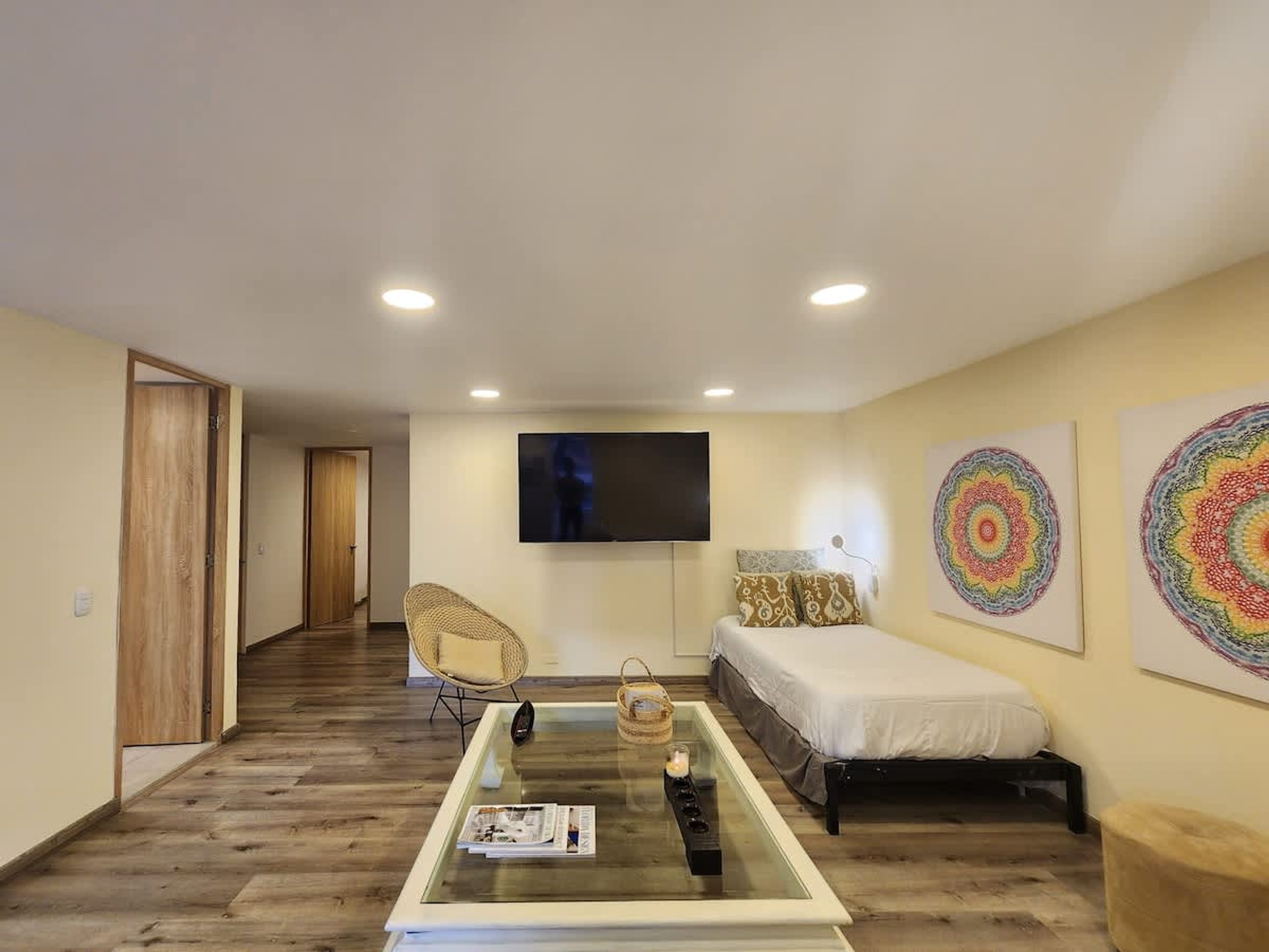 A modern hotel room with a glass coffee table, a bed, a TV, and two large decorative wall art pieces.