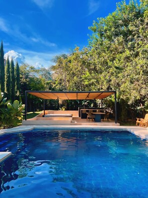 Una piscina con zona de estar cubierta, rodeada de abundante vegetación.