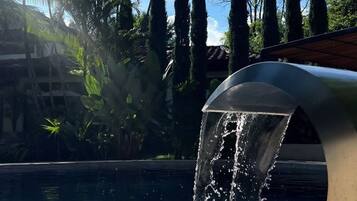A pool with a water feature, surrounded by palm trees and tall cypress trees.