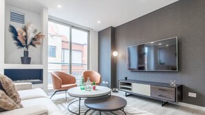 A modern living room with a sofa, two armchairs, a round coffee table, a flat-screen TV mounted on the wall, a sideboard, and a decorative plant. - Cozy 3 Br Apartment in Usaquen (Bogotá)