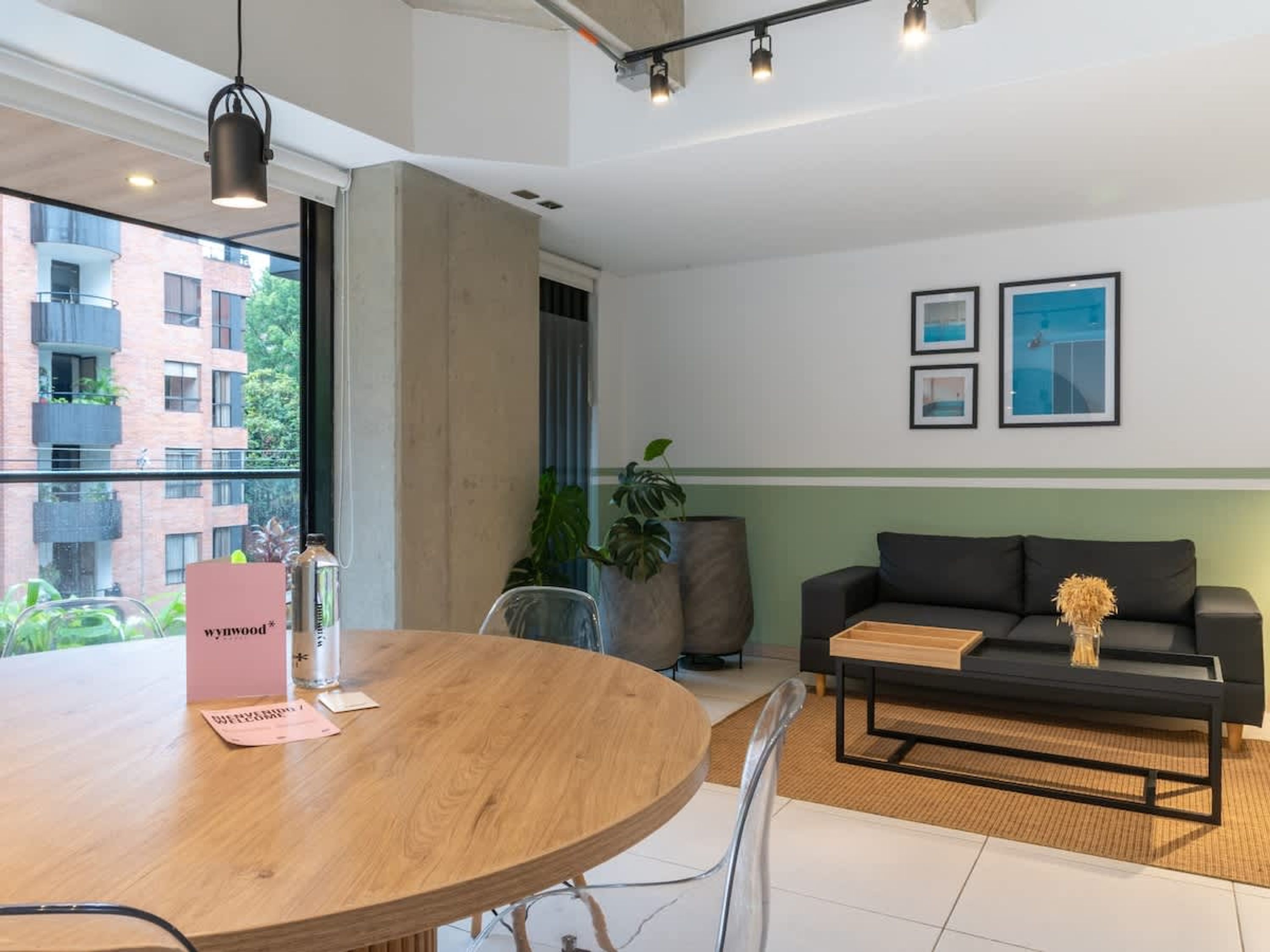 Un salon moderne avec une table ronde en bois, un canapé noir et une grande fenêtre donnant sur un immeuble et de la verdure.