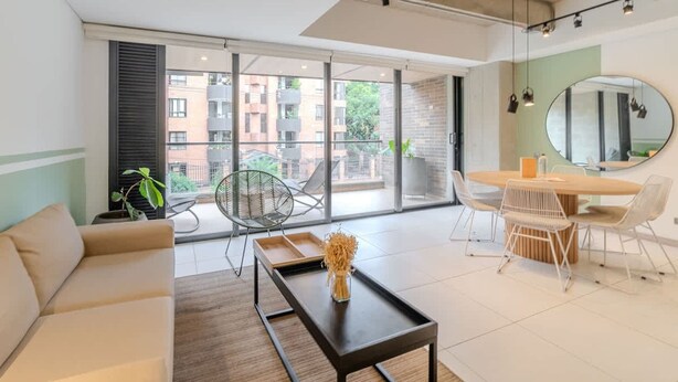 Een moderne woonkamer met een bank, een eettafel en stoelen. Er is een groot raam met uitzicht op gebouwen en groen.