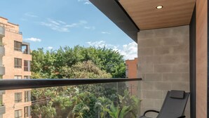 A balcony with a black lounge chair, a glass railing, and a view of a residential area with trees and buildings. - Stylish 3BR in Laureles MDE (Medellín)