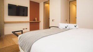 A modern hotel room with a large bed, a flat-screen TV mounted on the wall, a wooden shelf, and a door leading to another room. - Cozy 3 BR in Laureles in Medell n (Medellín)