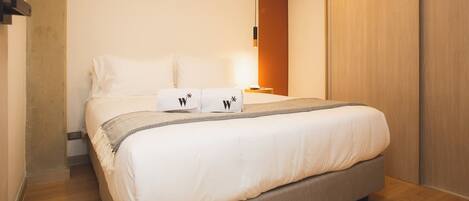 A neatly made bed with white linens and two pillows, a bedside table with a lamp, and a wooden wardrobe.