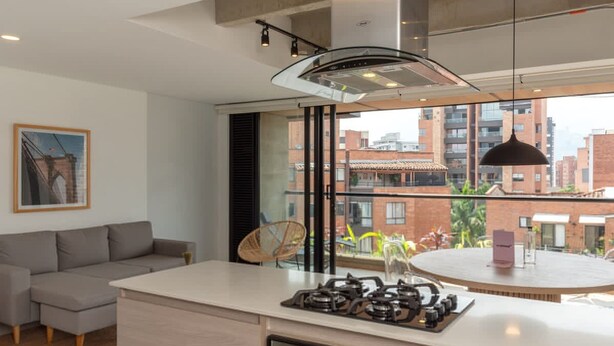 A modern kitchen with a central island, a dining area with a round table, and a view of the city through large windows. - Beautiful 3BR in Laureles Medell n (Medellín)